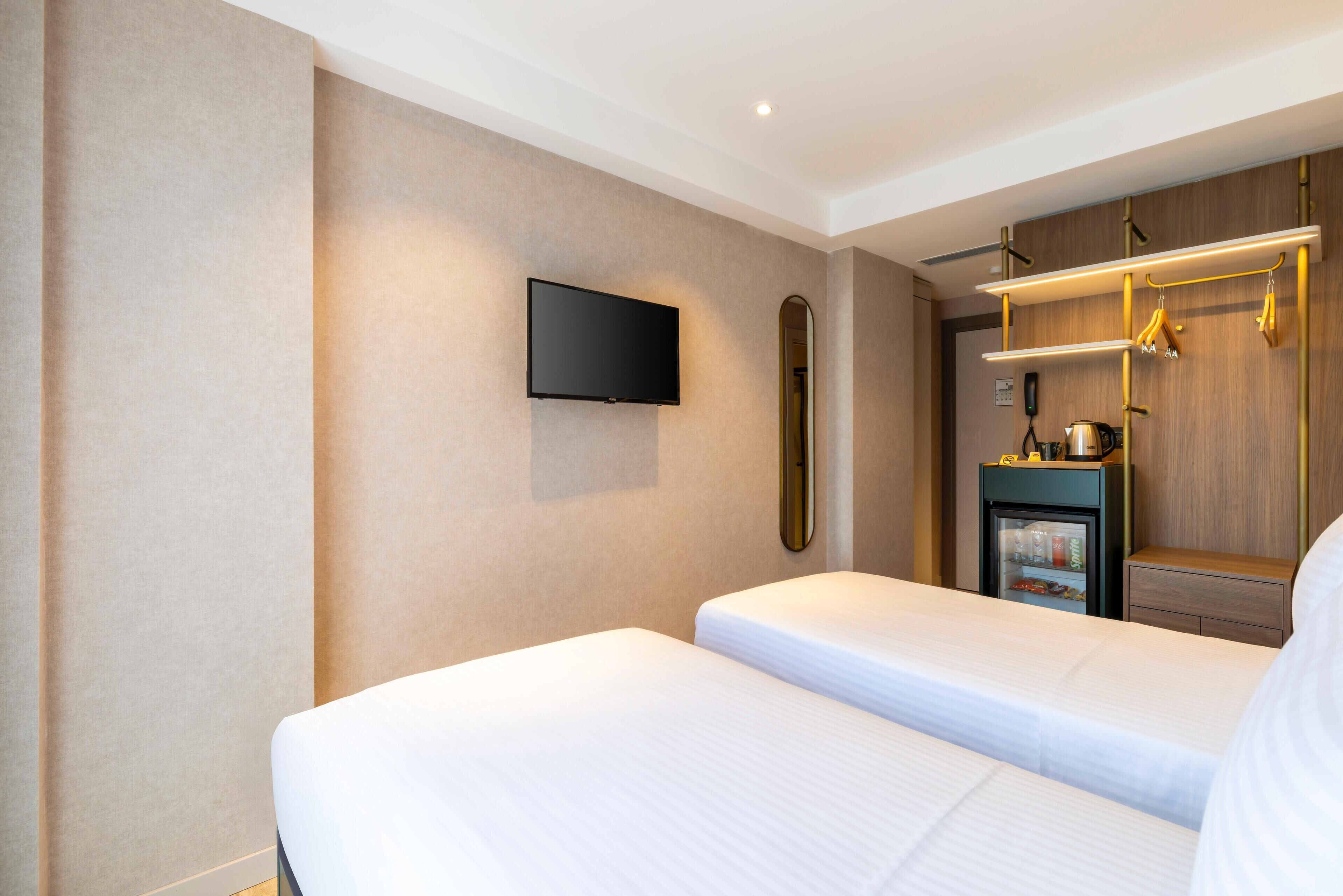 A hotel room with two beds, a television mounted on the wall, a minibar, and a wooden wardrobe with a mirror.