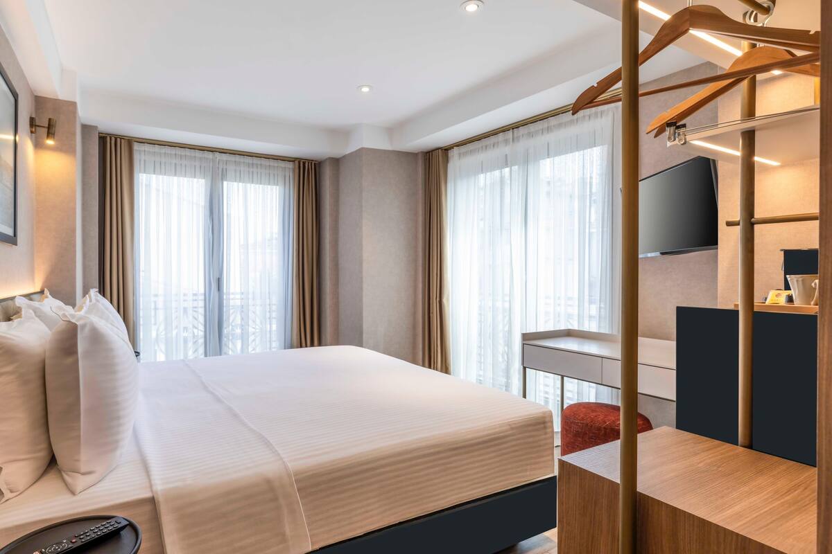 A hotel room with a large bed, a desk with a chair, a TV mounted on the wall, and a window with curtains.