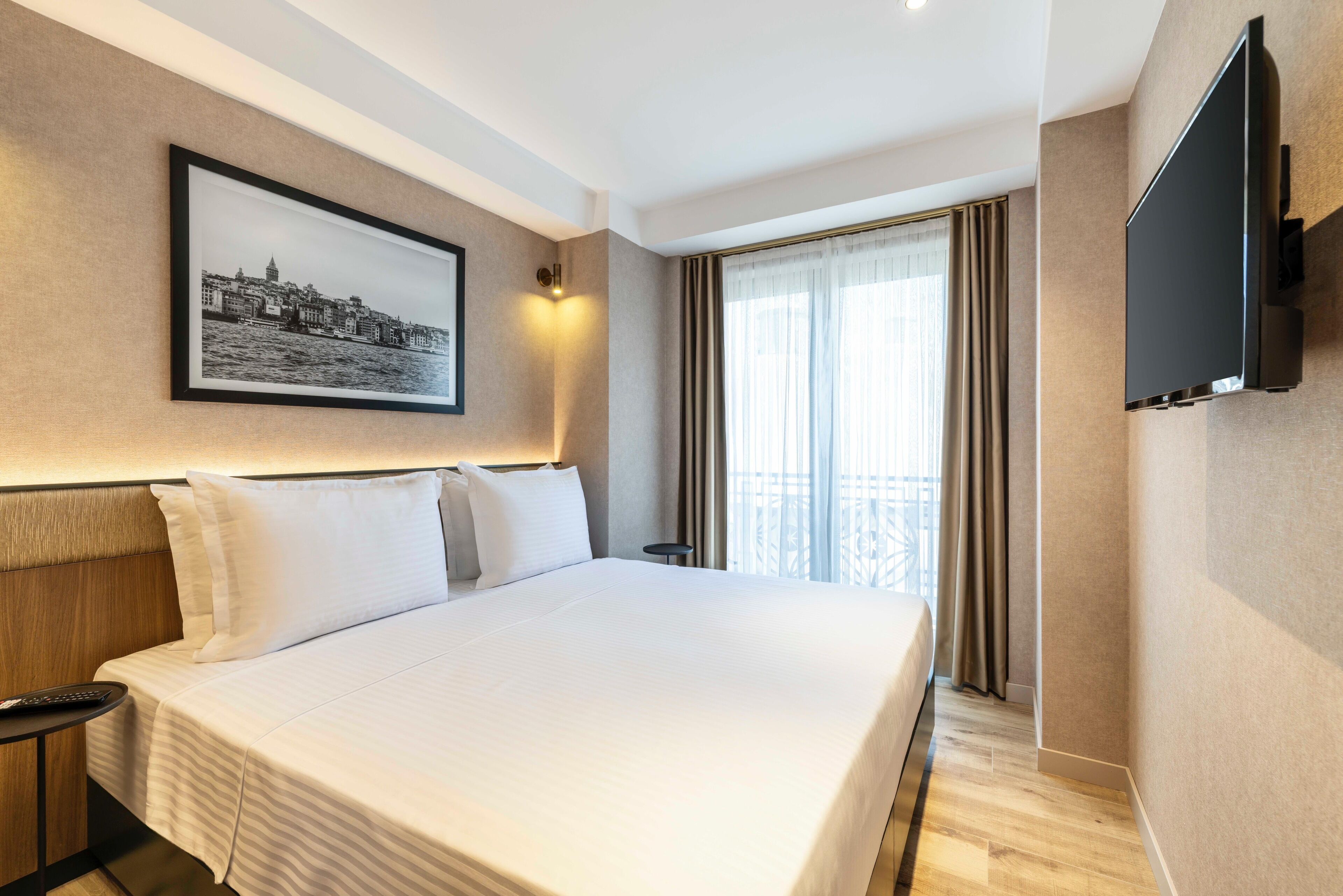 A hotel room with a large bed, a mounted TV, a framed cityscape picture, and a window with curtains.