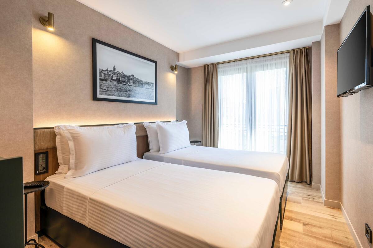 A hotel room with two beds, a mounted TV, a painting on the wall, and a window with curtains.