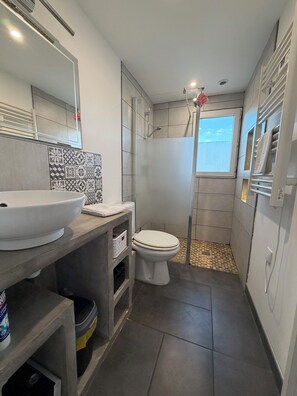 Une salle de bain avec un grand lavabo blanc, des toilettes, une douche avec une fenĂȘtre et des carreaux dĂ©coratifs sur le mur.