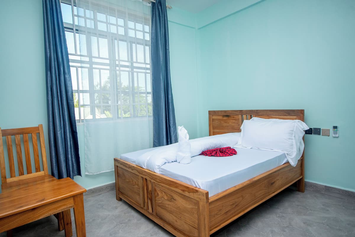 A single-bed room with a wooden headboard, a wooden chair, and a window with blue curtains.