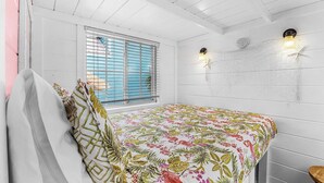 A room with a bed, a window with blinds, a wall-mounted light fixture, and a small stool.