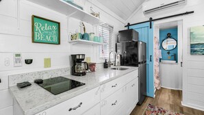 Une cuisine avec des meubles blancs, un réfrigérateur noir, une machine à café et une porte bleue donnant sur une salle de bain.