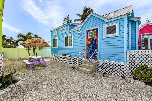 A blue house with a colorful mural on the door, a purple table, and a thatched umbrella. - Matlacha Tiny Village - Dolphin (Matlacha)