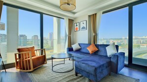 A modern living room with a blue sofa, a wooden chair, and a small round table. Large windows offer a city view. - Modern Furnished Apartments in Istanbul Basaksehir By Iradah Gayrimenkul (Istanbul)