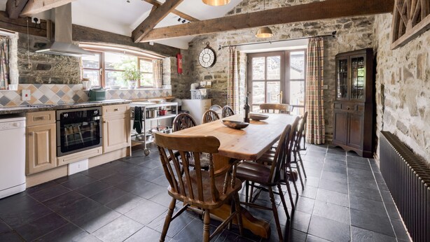 A rustic kitchen with exposed beams, a stone wall, and a large wooden dining table with chairs. - Coach House (Llandysul)