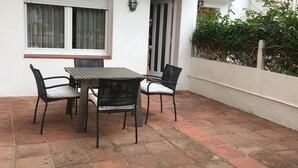 Une terrasse avec une table et quatre chaises, une fenêtre avec des rideaux, un luminaire fixé au mur et un petit balcon agrémenté de plantes.