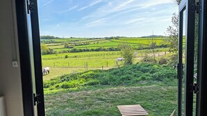 Une vue depuis une pièce dotée d’une porte-fenêtre coulissante, qui dévoile un paysage rural avec des collines ondoyantes, des champs verdoyants et quelques chevaux qui paissent.