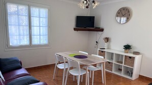 En stue med en rød lædersofa, et hvidt spisebord med fire stole, et fjernsyn på en træhylle, et ur på væggen og et hvidt konsolbord med kurve.