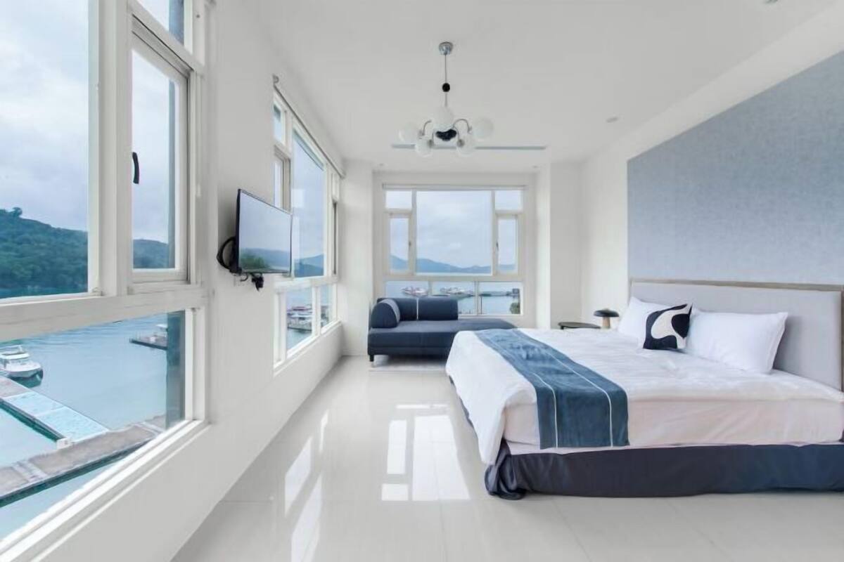 A hotel room with a bed, a TV, a balcony with a view of the sea, and a large window.