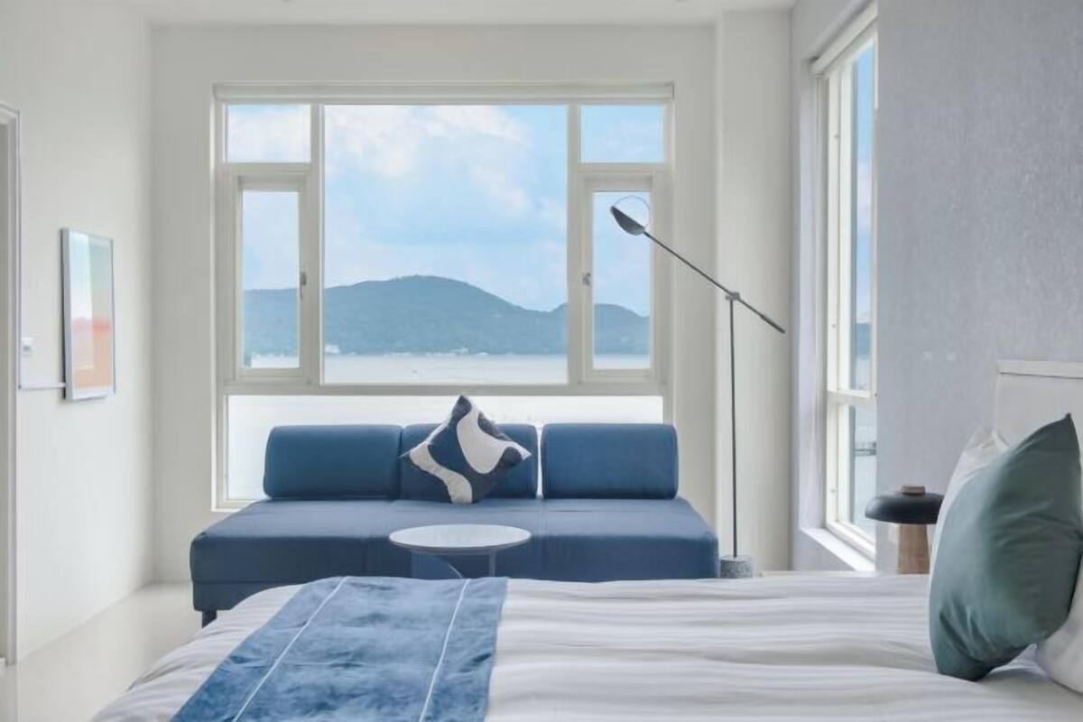 A bedroom with a bed, a blue sofa, a round table, and a large window with a view of the sea and mountains.