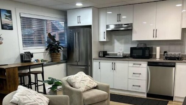 Cocina compacta con alacenas blancas, refrigerador, microondas y pileta. Hay dos sillones beige con cojines, una pequeña mesa de comedor y una planta en maceta.
