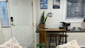 Una habitación con dos sillones color beige, una mesita redonda con una planta en maceta, una barra de madera y una ventana con persianas.