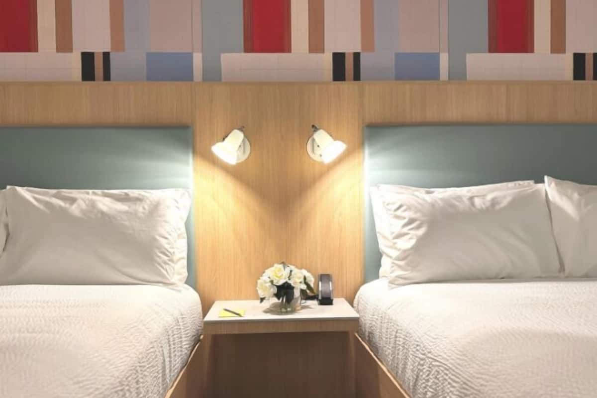 A hotel room with two beds, a wooden headboard, bedside lamps, a nightstand with a vase of flowers, and a wall with abstract art.