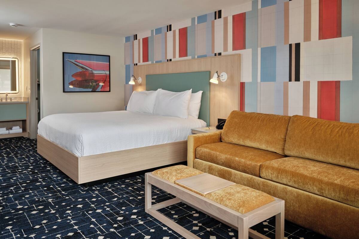A hotel room with a bed, a sofa, a small table, a nightstand, a wall with a colorful abstract design, and a painting of a red boat.