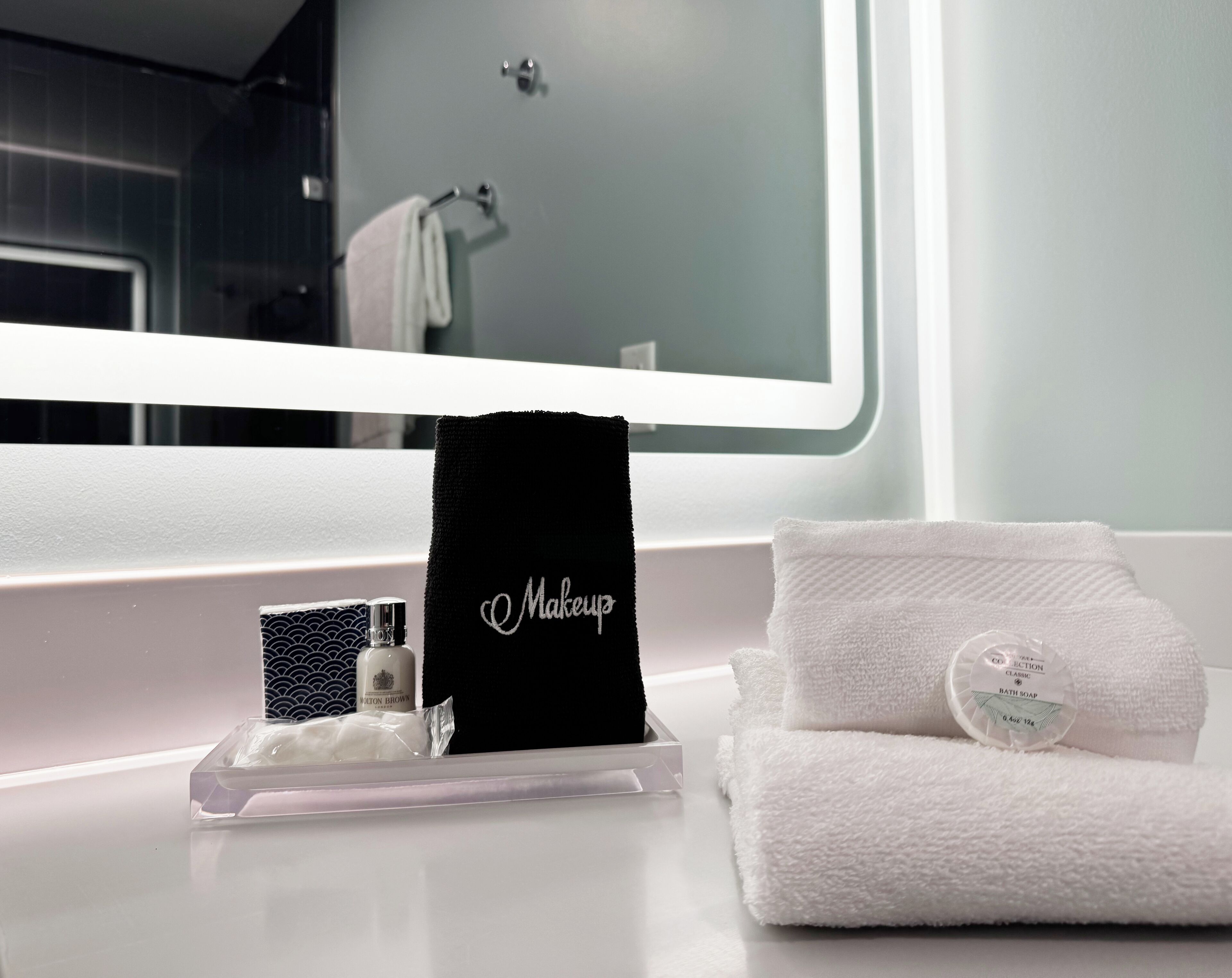 A bathroom vanity with a mirror, a black towel with 'Makeup' embroidered, a white towel, and a bottle of bath soap.
