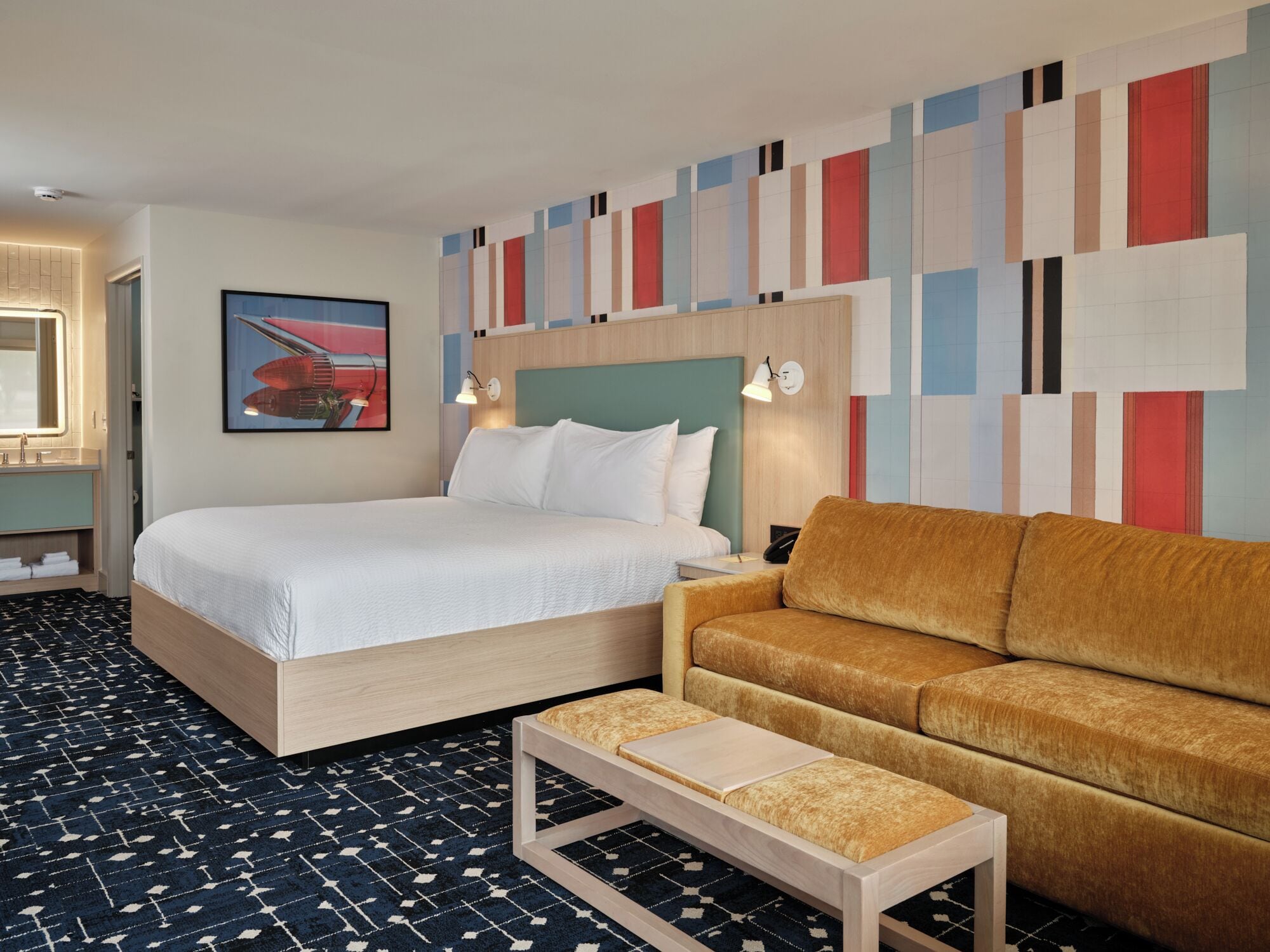 Una habitación de hotel con una cama, un sofá, una mesita pequeña, una mesita de noche, una pared con un diseño abstracto y colorido, y un cuadro de un bote rojo.