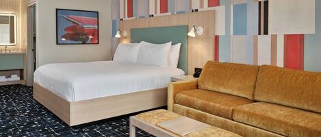 Una habitación de hotel con una cama, un sofá, una mesita pequeña, una mesita de noche, una pared con un diseño abstracto y colorido, y un cuadro de un bote rojo.