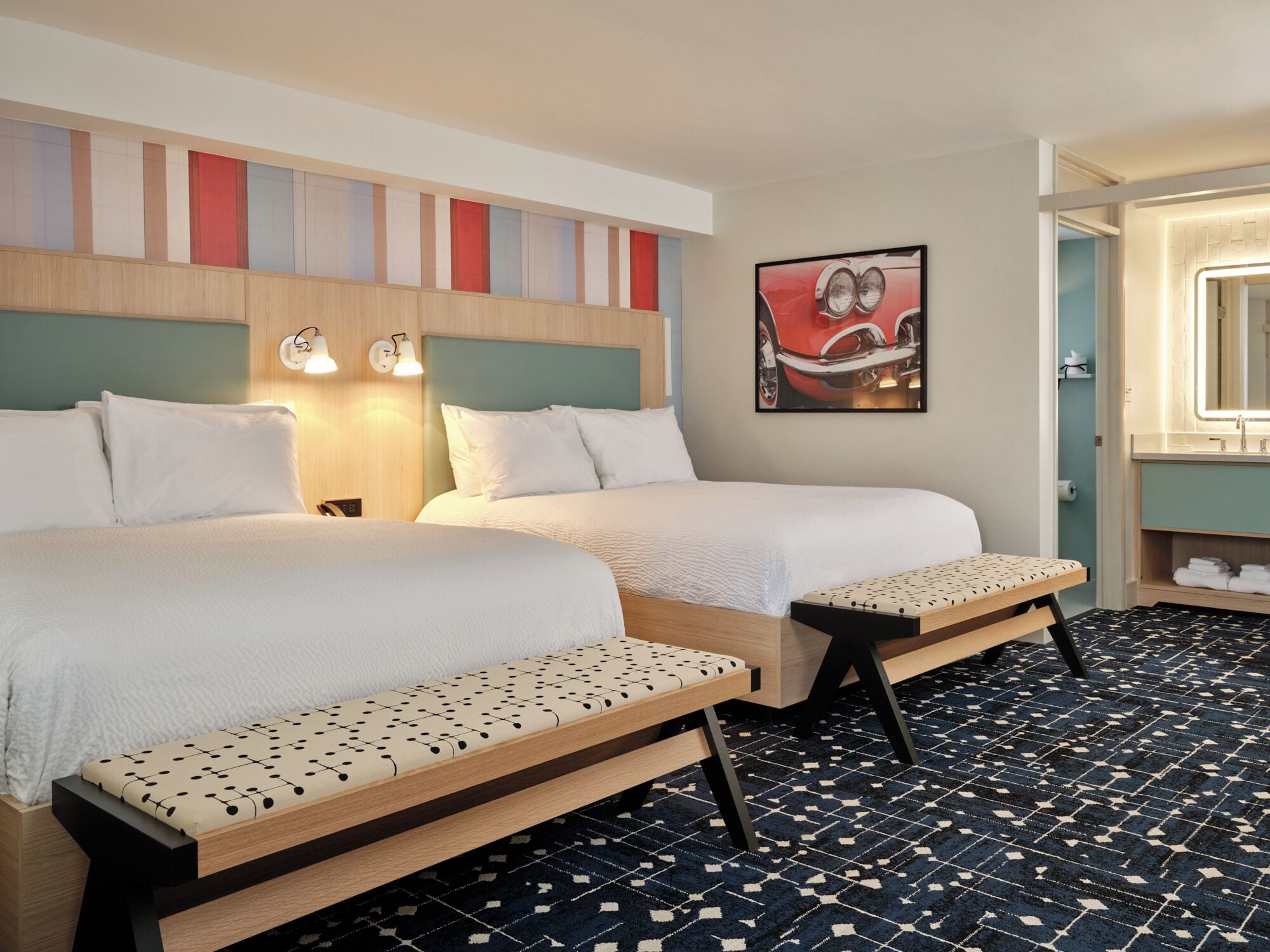 A hotel room with two beds, a bench, a painting, and a bathroom with a mirror.