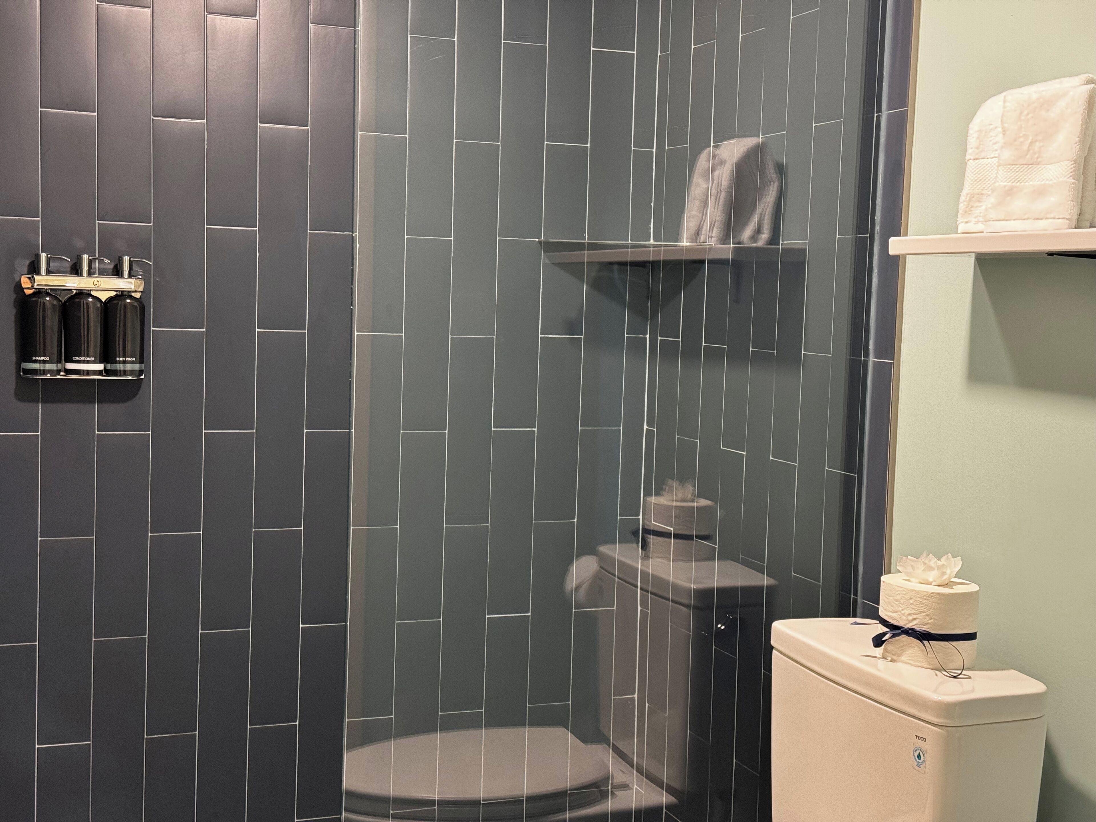 Une salle de bain au design moderne, avec des murs recouverts de carreaux foncés, des toilettes, une douche et une étagère contenant des articles de toilette.