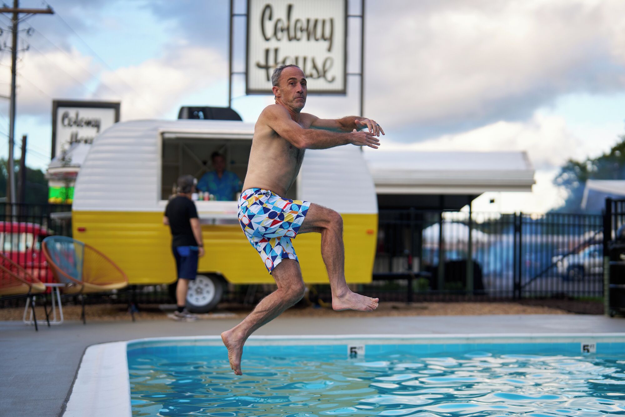 Ein Mann springt in einen Pool, im Hintergrund ein Schild mit der Aufschrift 'Colony House'.