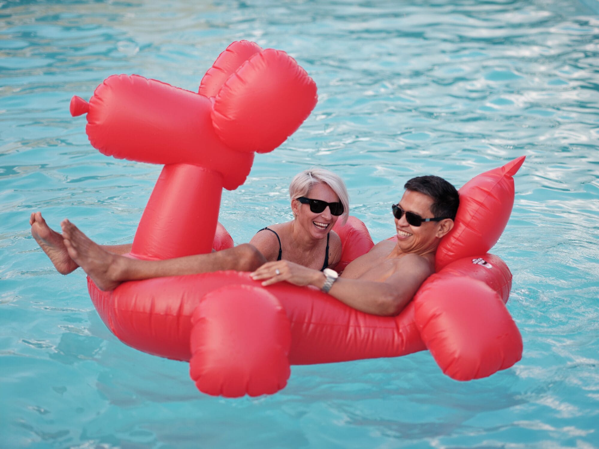 Two people floating in a pool using red inflatable floats shaped like a bear.