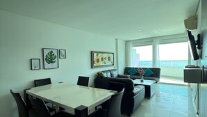 A modern living room with a dining area, featuring a white table, black chairs, a sofa, and a coffee table. There are framed pictures on the wall and a large window providing a view of the outdoors. - APARTAMENTO 702A (Cartagena)
