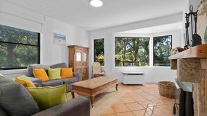 Un salón amplio con un sofá gris, una mesa de centro de madera y vistas al exterior a través de amplias ventanas.