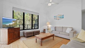 Una sala de estar con televisión, muebles de madera, un sofá y un ventanal con vista a árboles.