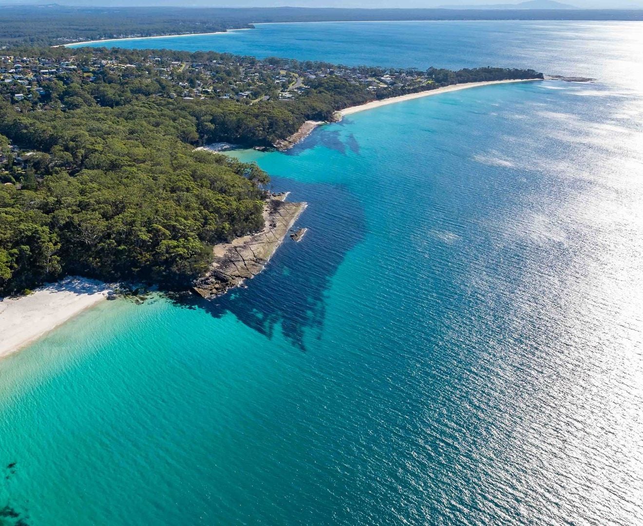 An aerial view of Jervis Bay with a beach, clear turquoise waters, and dense forested areas.
