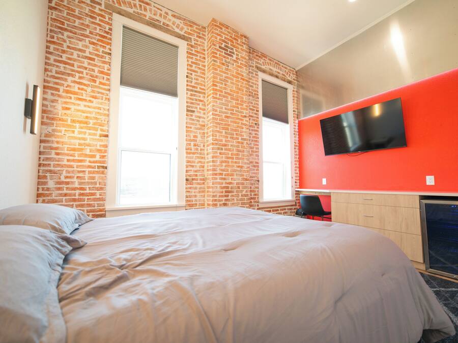 Una habitación de hotel con pared de ladrillo, una cama grande, un televisor de pantalla plana montado en la pared y una ventana con persianas.
