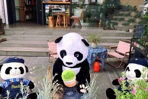 Une mascotte panda tenant un objet vert, assise sur une chaise, avec une table et d’autres chaises en arrière-plan.