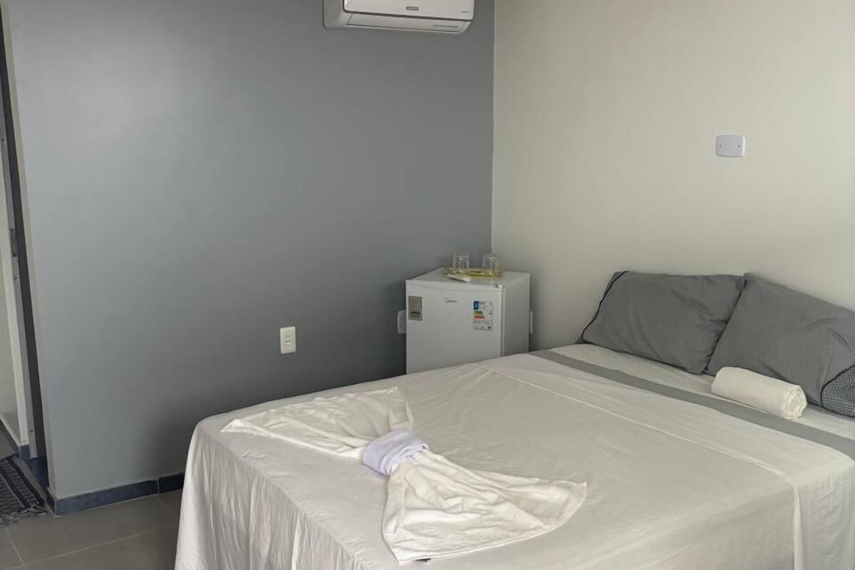 Une chambre d’hôtel avec un grand lit, une petite table de chevet, un climatiseur fixé au mur et un mur de couleur grise.