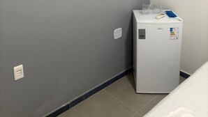 A small room with a white air conditioner mounted on the wall, a white refrigerator with a sticker, and a bed with white bedding. - Hospedagem Praia do Forte (Tamandaré)