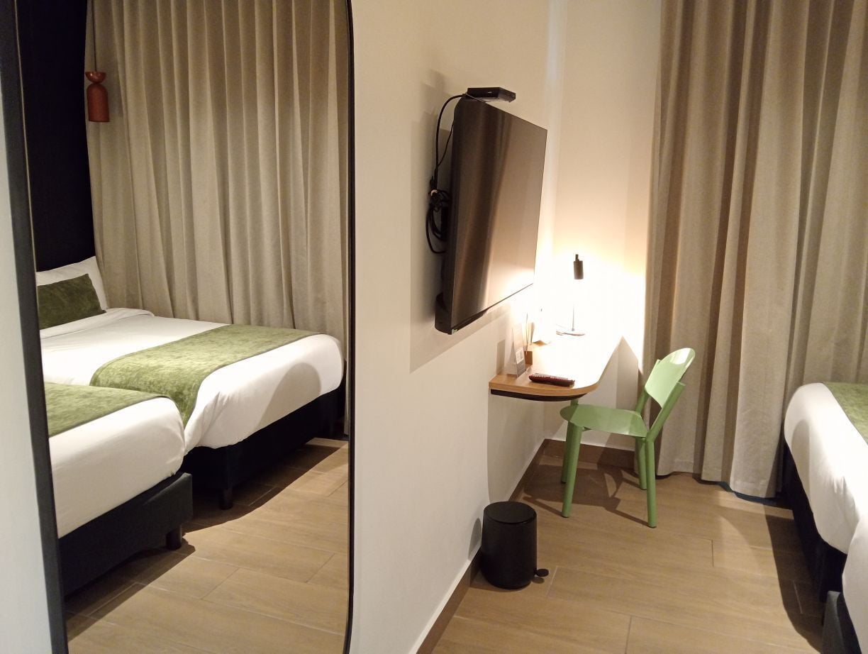 Une chambre d’hôtel avec deux lits, un bureau avec une chaise, un téléviseur fixé au mur et une petite poubelle.
