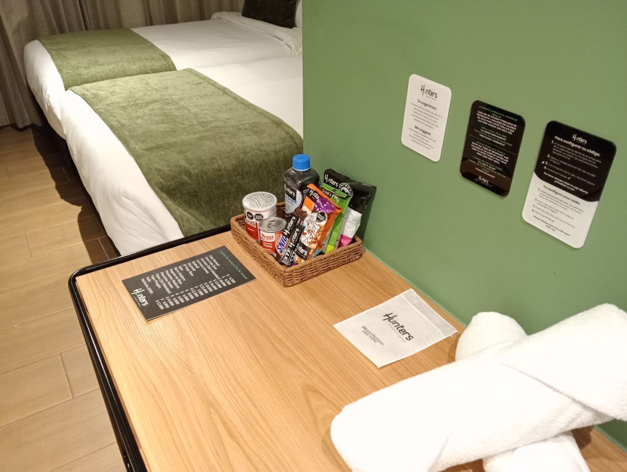 A hotel room with a bed, a small table with a menu, a basket of snacks, and a green wall with informational cards.