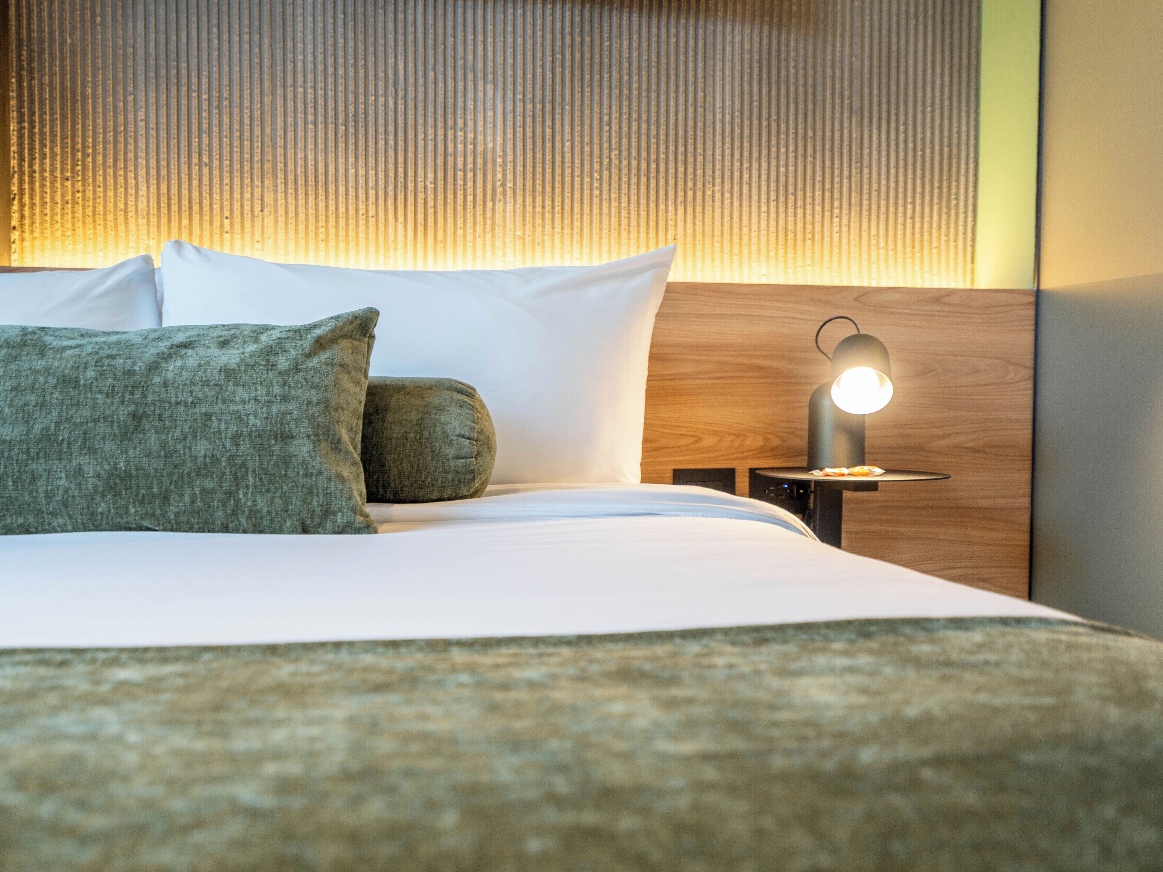 Una cama bien hecha con almohadas, una mesita de noche con lámpara y un cabecero de madera.