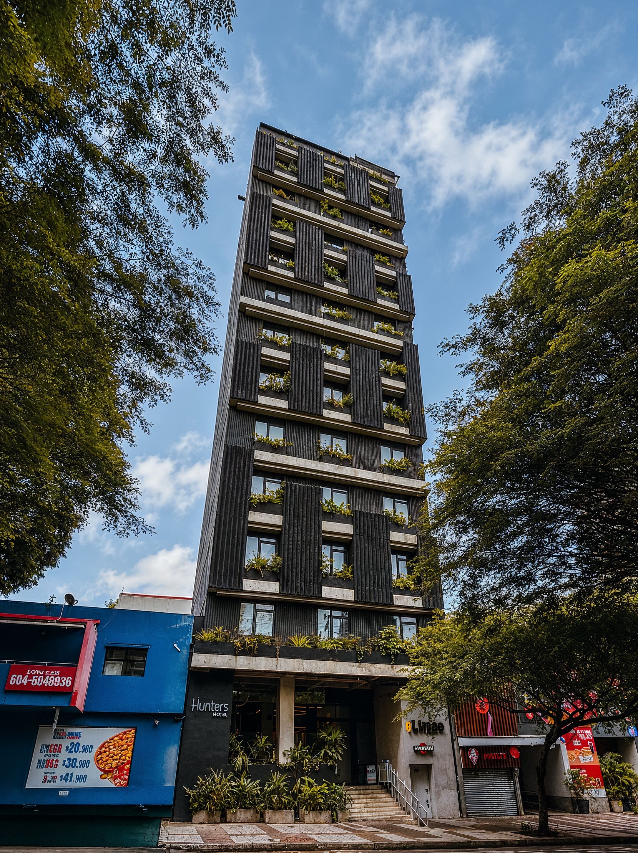 Un edificio moderno con varios pisos, balcones con vegetación y una entrada visible.