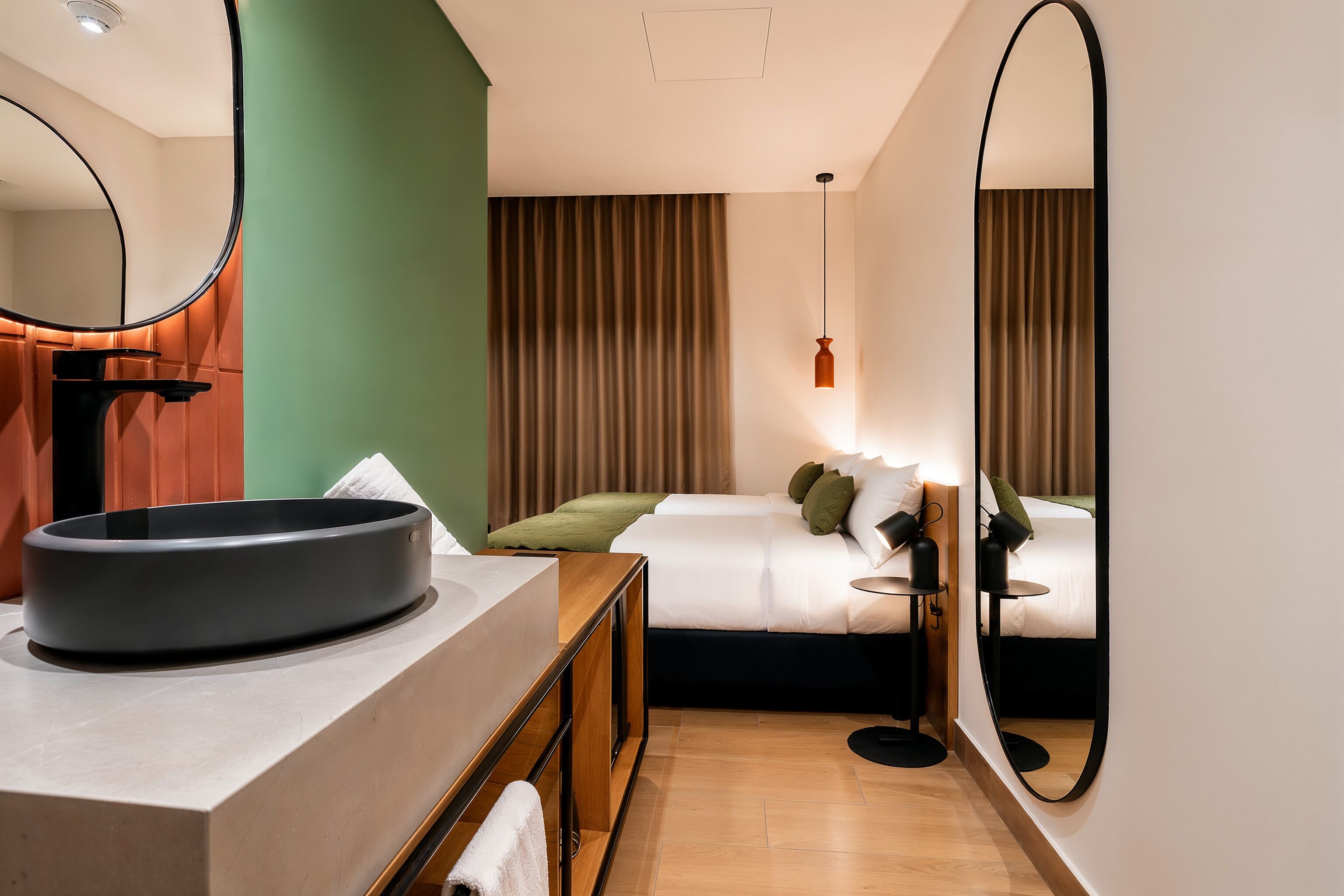 A hotel room with a round sink, a bed with pillows, a bedside table, and a large mirror.