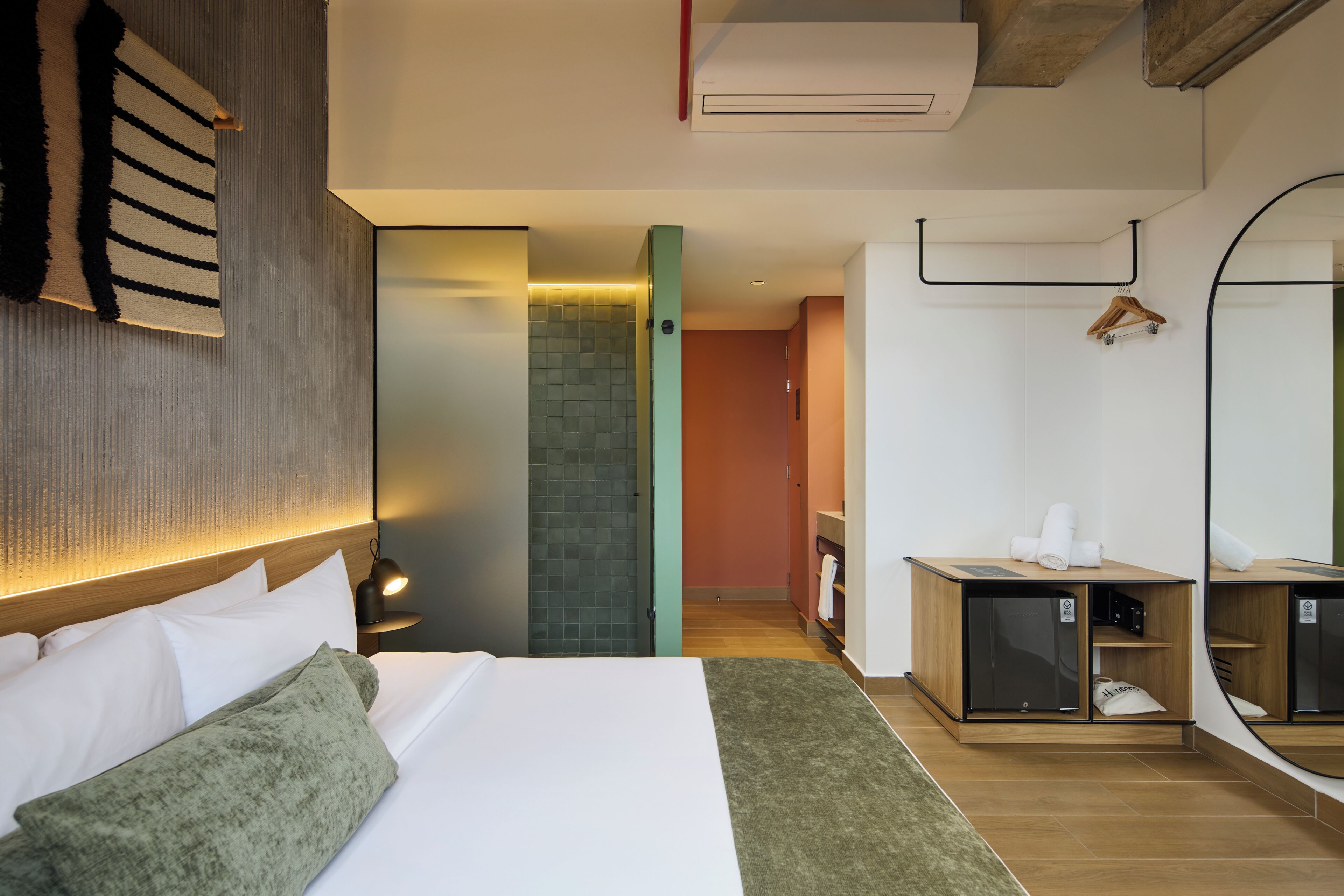 Une chambre d’hôtel avec un lit, une table de chevet, un miroir, une armoire en bois et une salle de bain visible à travers une porte ouverte.