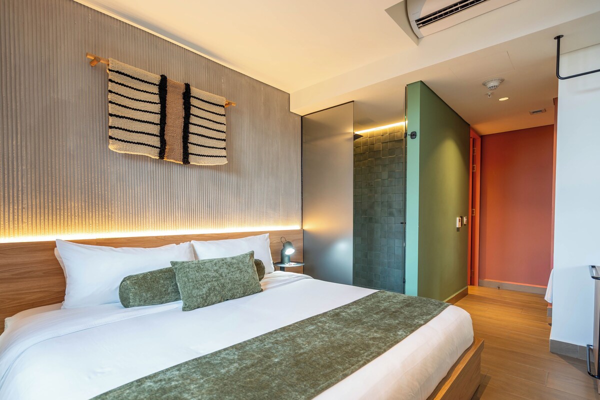 Ein modernes Hotelzimmer mit einem großen Bett, einem Nachttisch mit Lampe, einer Wand-Klimaanlage und einer Schiebetür zu einem Badezimmer.