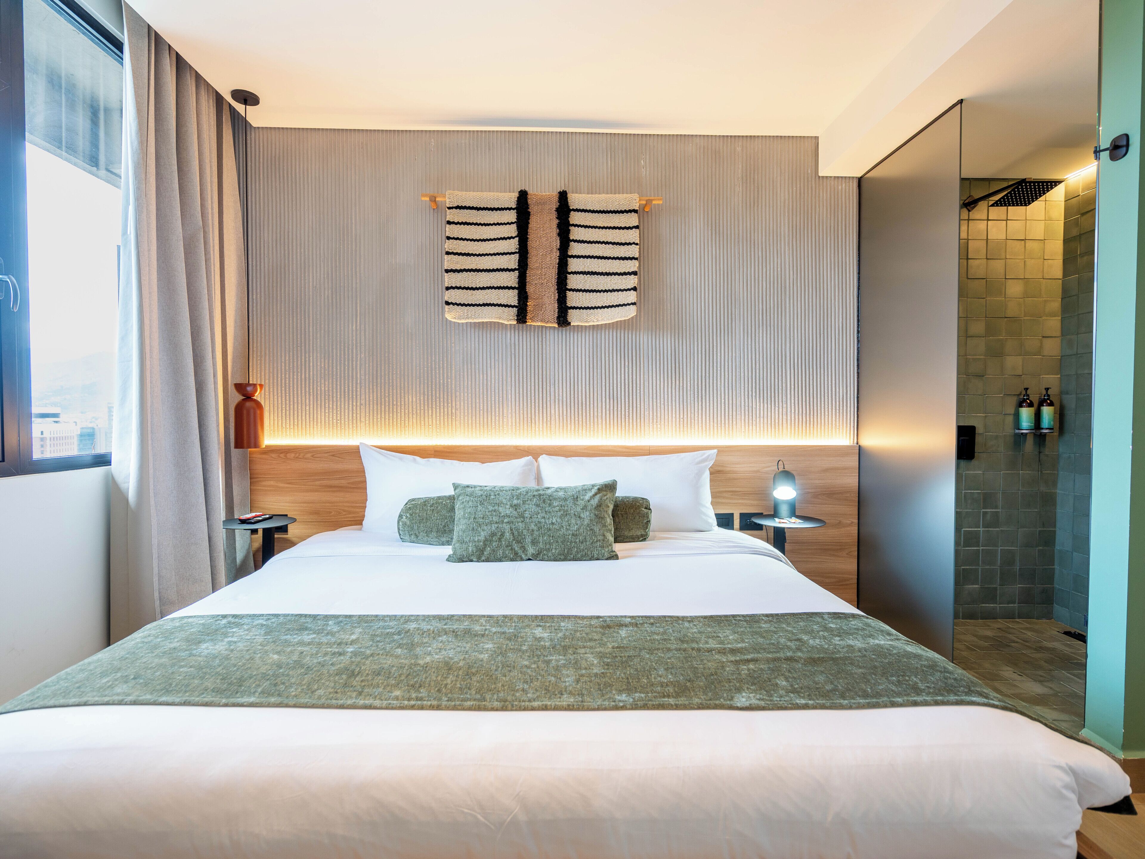 A modern hotel room with a large bed, a headboard with vertical lines, a wall-mounted towel rack, a nightstand with a lamp, and a view of the city through the window.