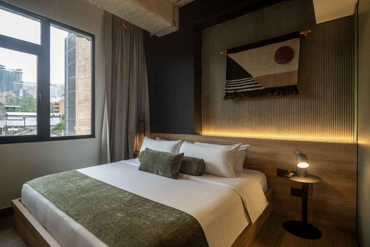 A hotel room with a large bed, a nightstand with a lamp, a view of the city through a window, and a wall-mounted tapestry.