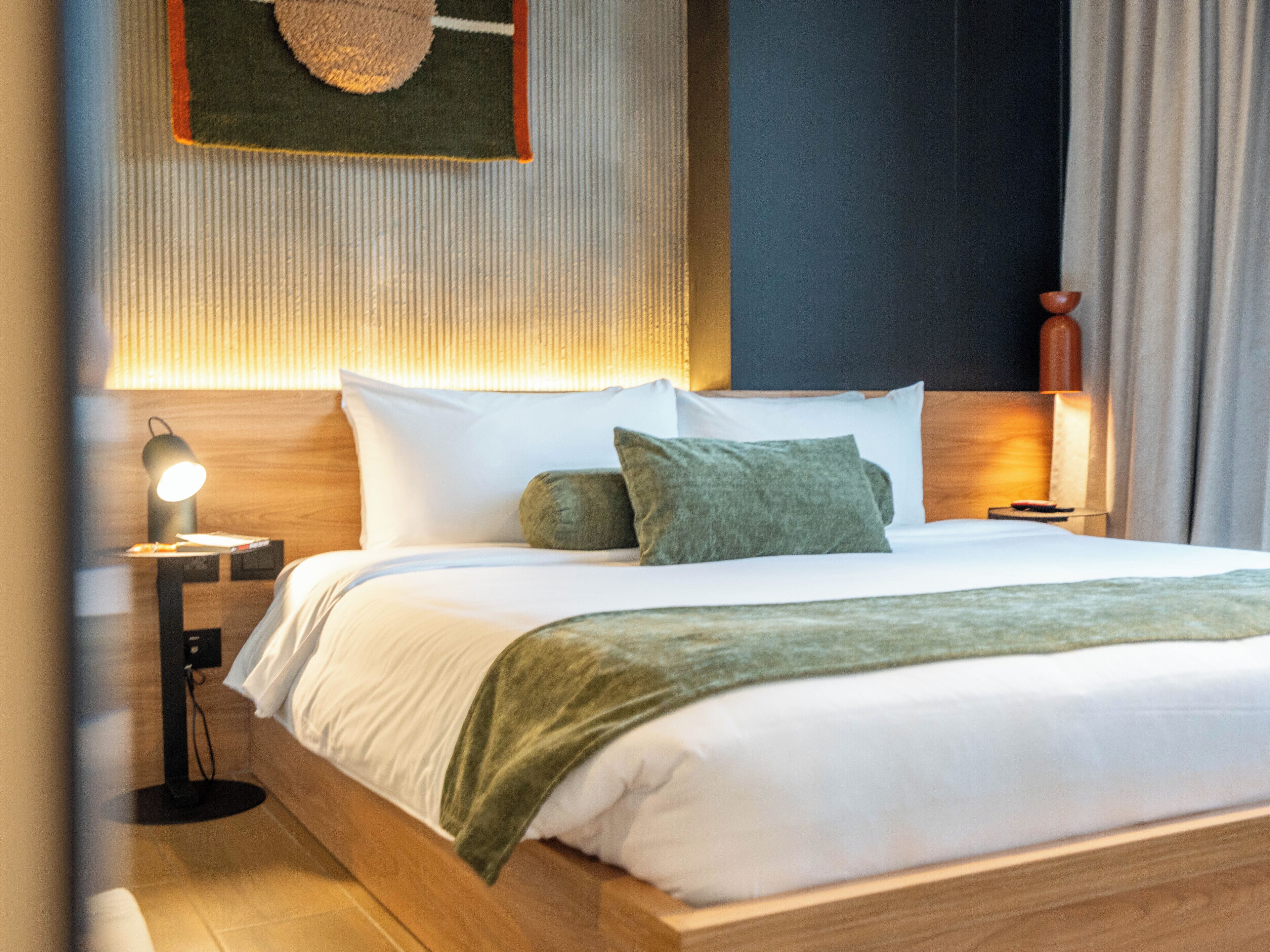 A neatly made bed with white linens and a green throw blanket, a bedside table with a lamp, and a wall-mounted light fixture.