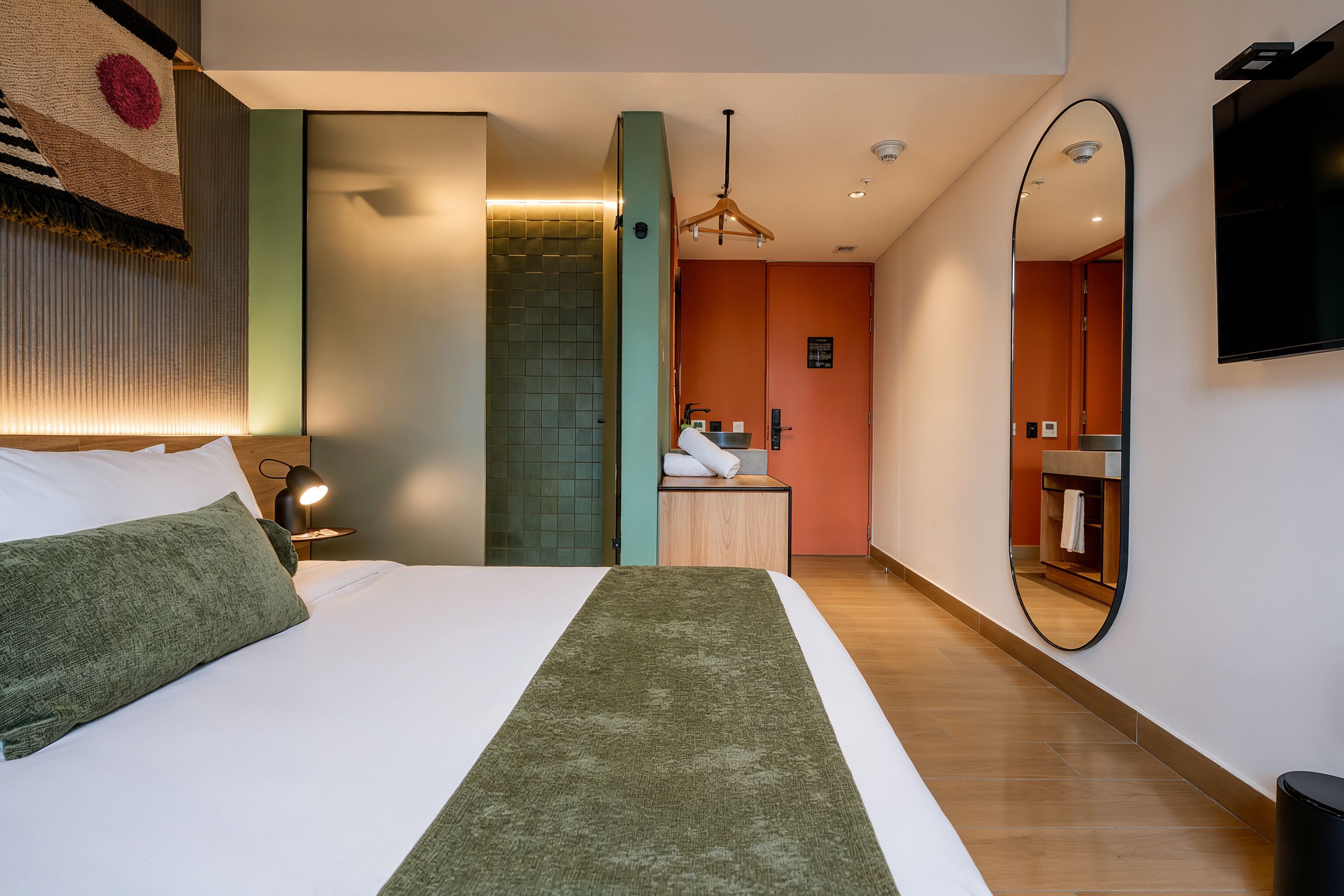 Une chambre d’hôtel avec un lit, une lampe de chevet, un miroir, une salle de bain avec un lavabo et un téléviseur fixé au mur.