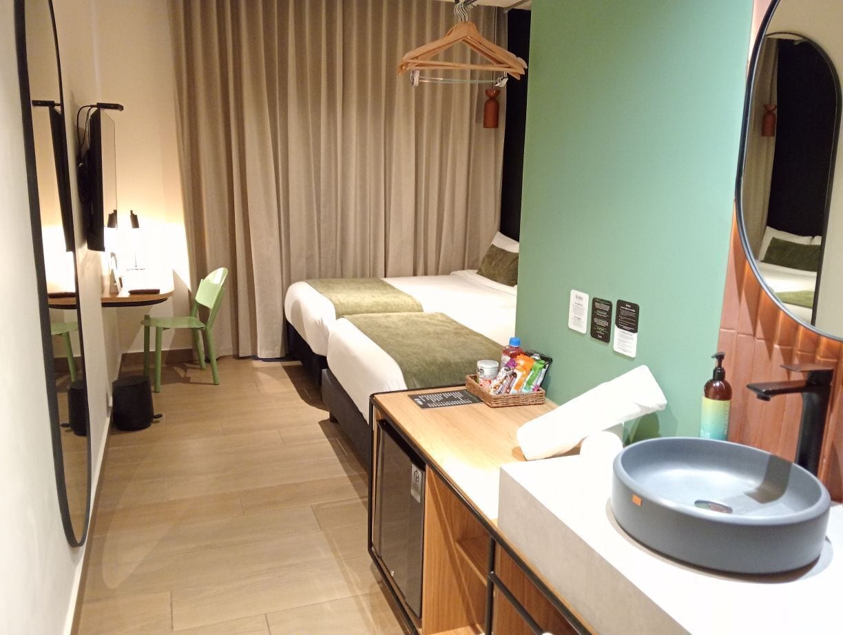 Habitación de hotel con cama, escritorio y silla, televisor pequeño, lavamanos y espejo.