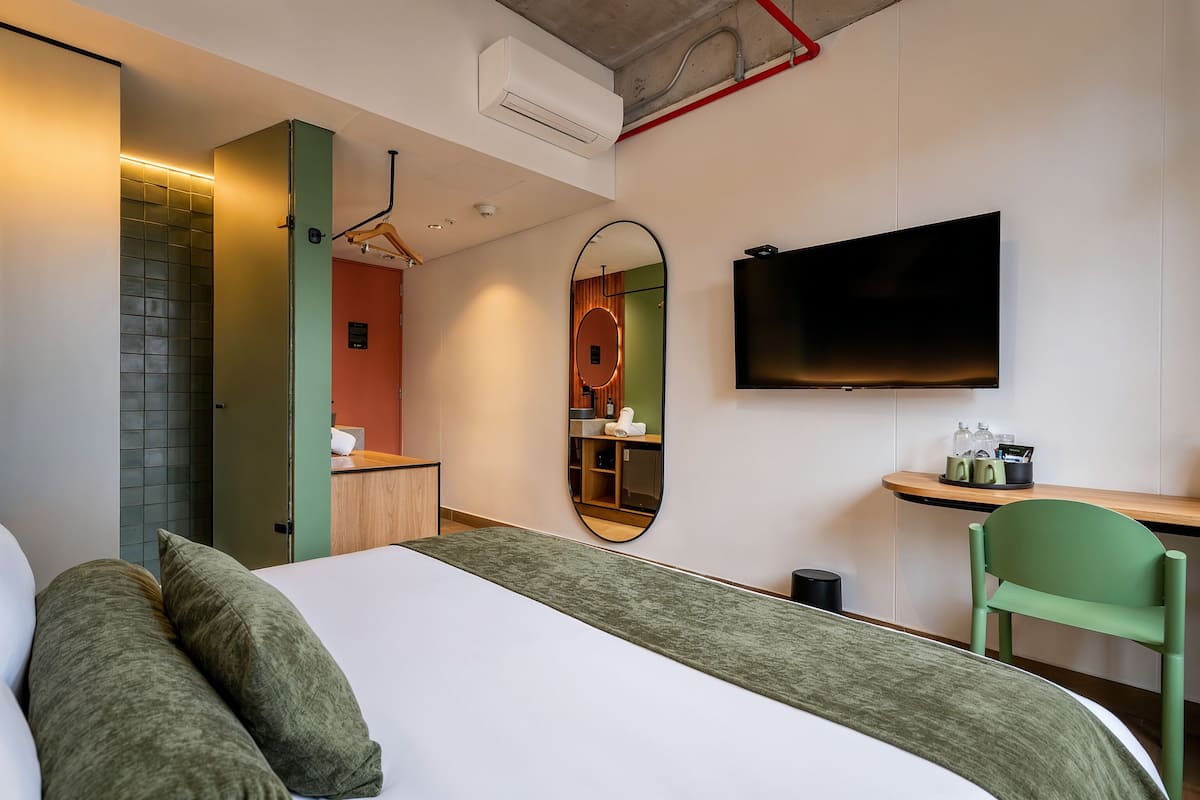 A hotel room with a bed, a green chair, a TV, a mirror, and a small desk with a shelf.