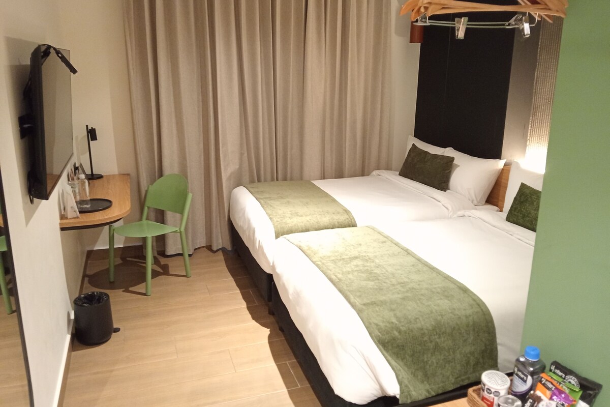 Hotellihuone, jossa on sänky, työpöytä, tuoli, seinälle kiinnitetty naulakko ja pieni pöytä, jonka päällä on joitain esineitä.