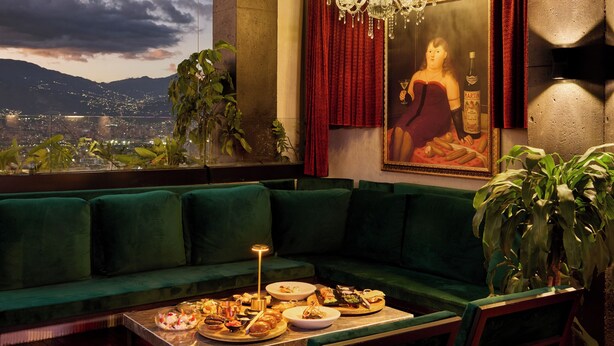 Una stanza con un divano verde, un tavolo da pranzo con piatti e posate, un lampadario e un'ampia finestra che offre vista sulla città e sulle montagne.